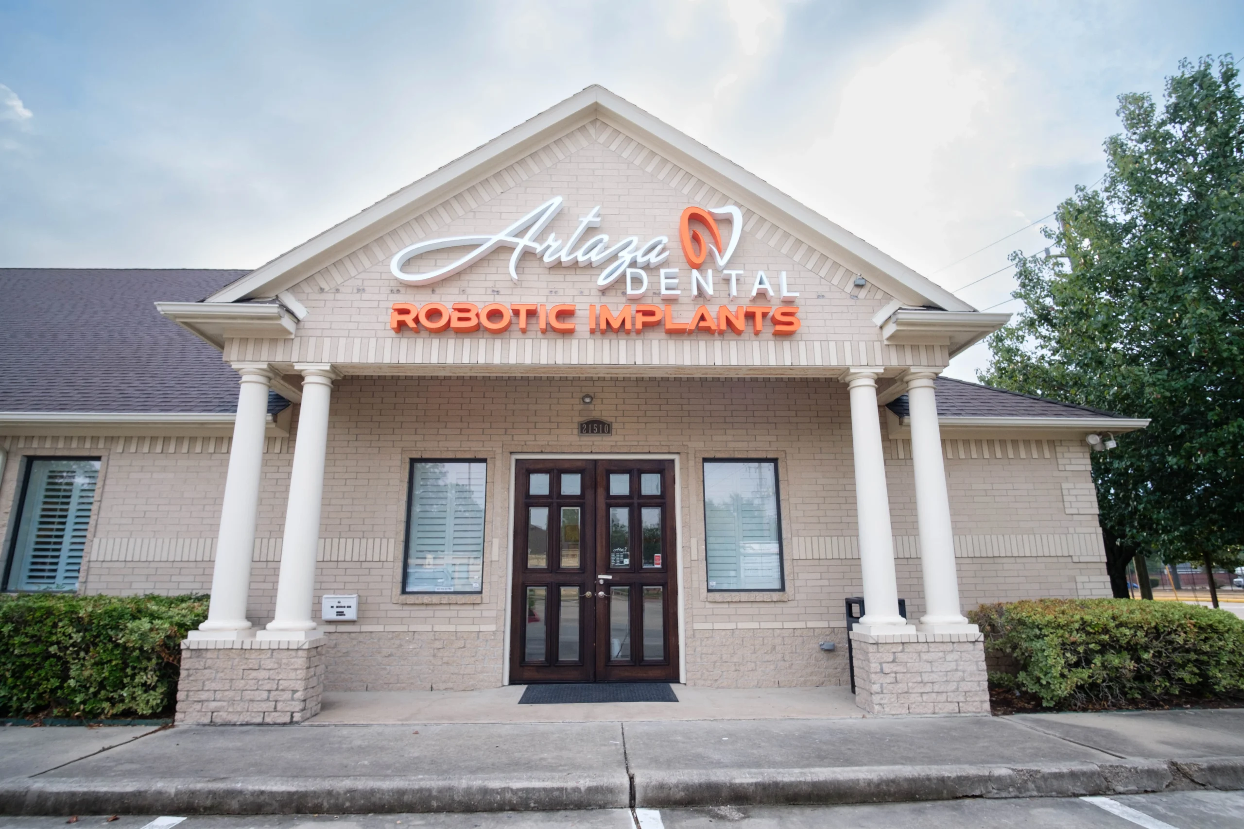 A dental office with a sign reading "Artaza Dental - Robotic Implants" is pictured. The building has a brick exterior, double glass doors, and white columns supporting a triangular pediment. There is a tree on the right side of the image.