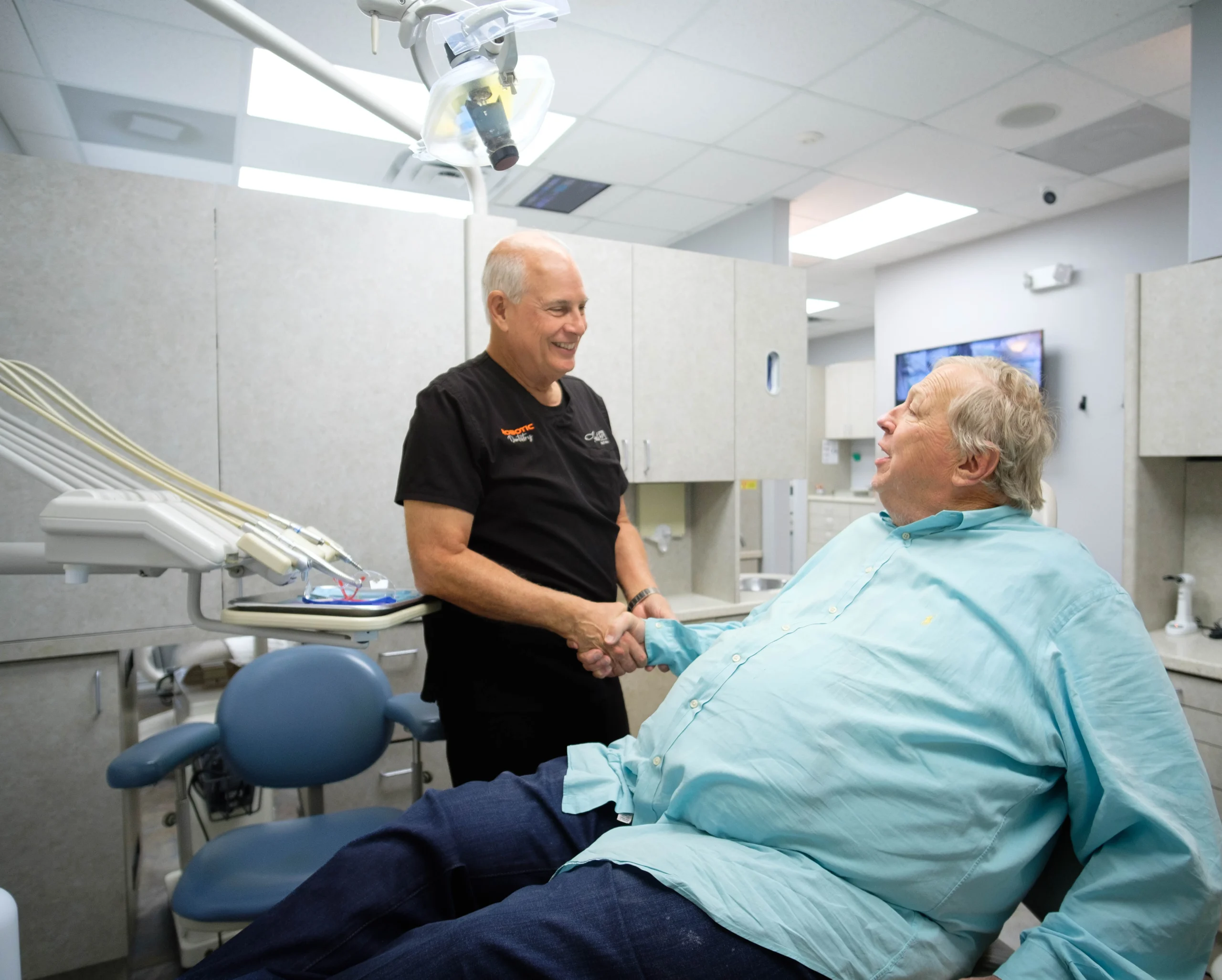 A dentist in black scrubs shakes hands with a man in a light blue shirt who is sitting in a dental chair. The room contains typical dental equipment and a monitor on the wall. Both individuals are smiling.