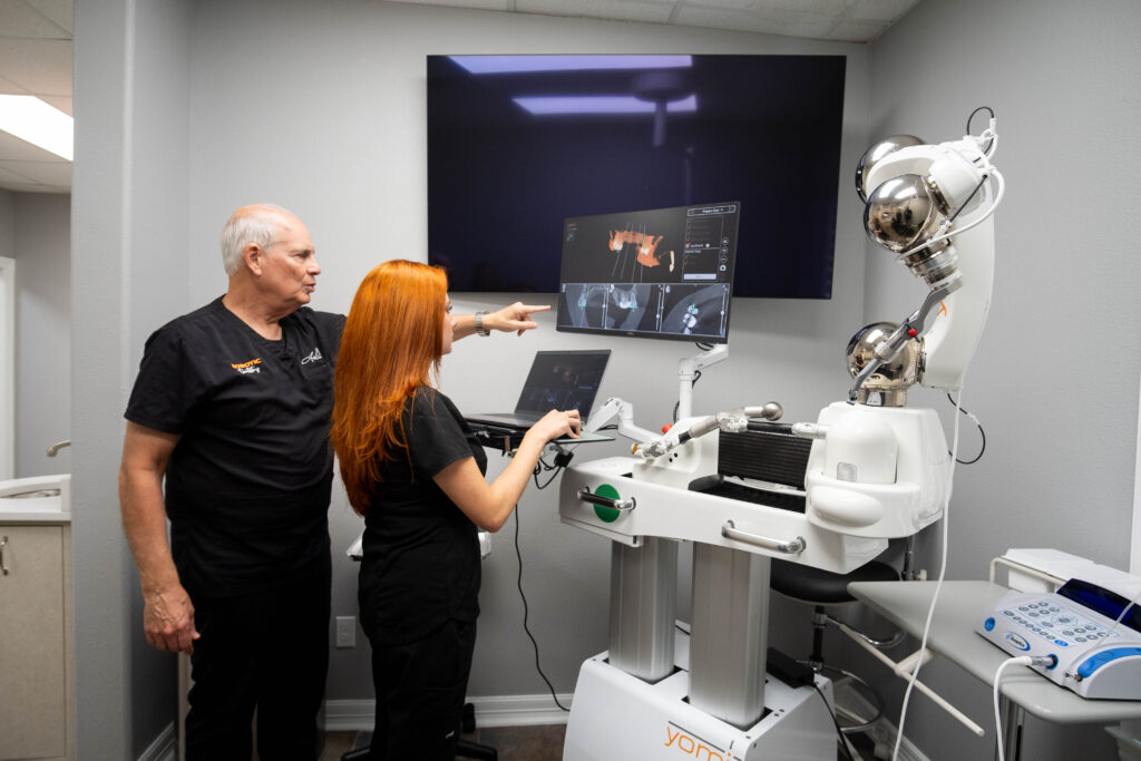 Two dental professionals in a clinic examine a monitor displaying dental images. One points at the screen while the other operates a computer. Nearby, a robotic device is present. Both wear black scrubs, and the room has a clinical setup with various medical equipment.