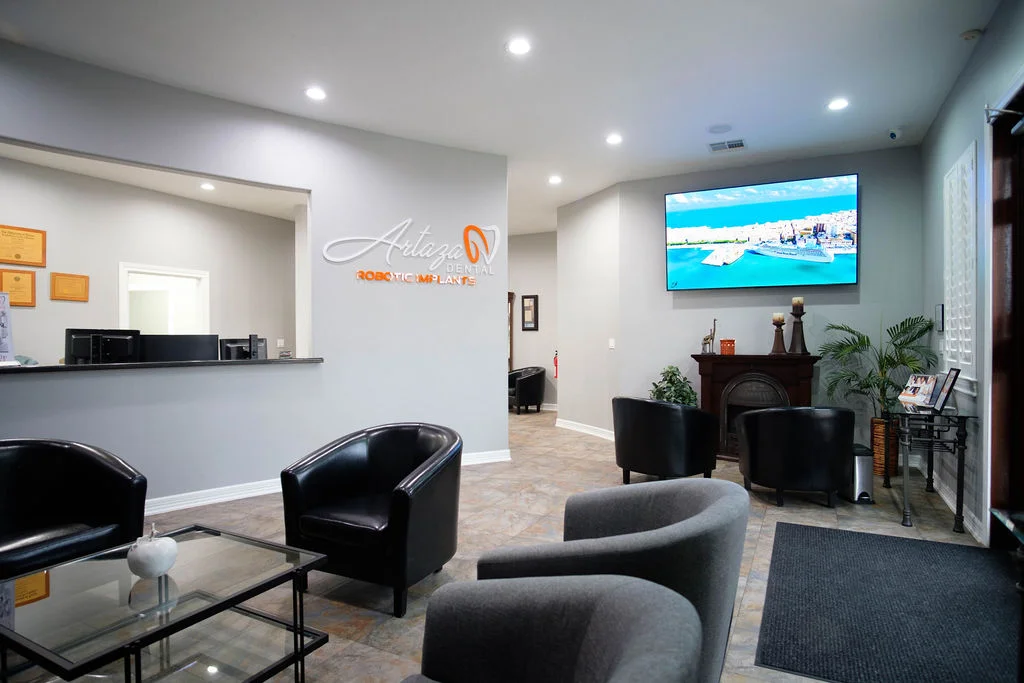 A modern dental office waiting area with four black chairs arranged around a glass coffee table. The room has a light grey color scheme, a wall-mounted TV displaying a beach scene, and framed certificates on the reception desk wall. The office is clean and well-lit.