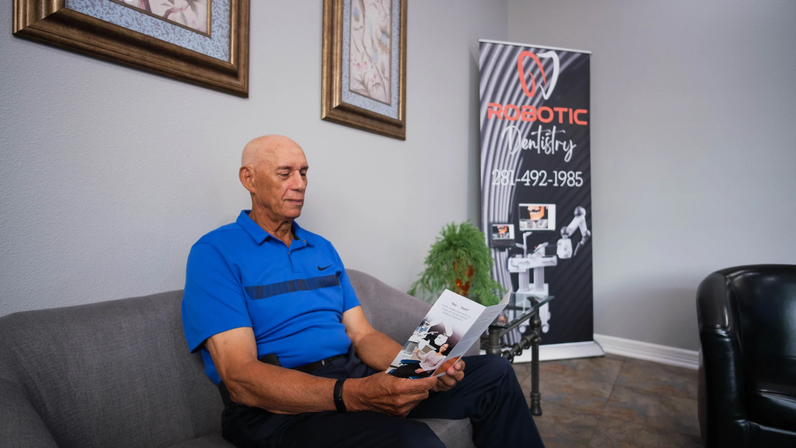 An elderly man in a blue polo shirt sits on a gray couch, reading a pamphlet. Behind him, there is a banner with the text "Robotic Ministry" and a phone number. The room has framed artwork on the walls and contemporary furniture.