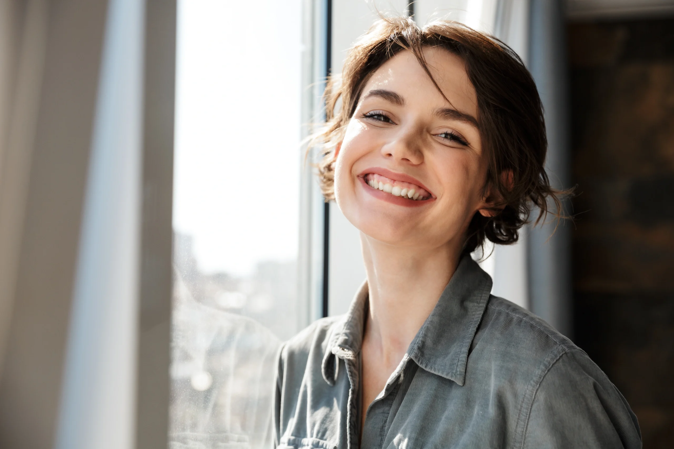A young woman with short brown hair and a casual grey shirt smiles warmly while standing near a window with natural light streaming in. The background shows an out-of-focus cityscape, adding a pleasant urban atmosphere to the image.