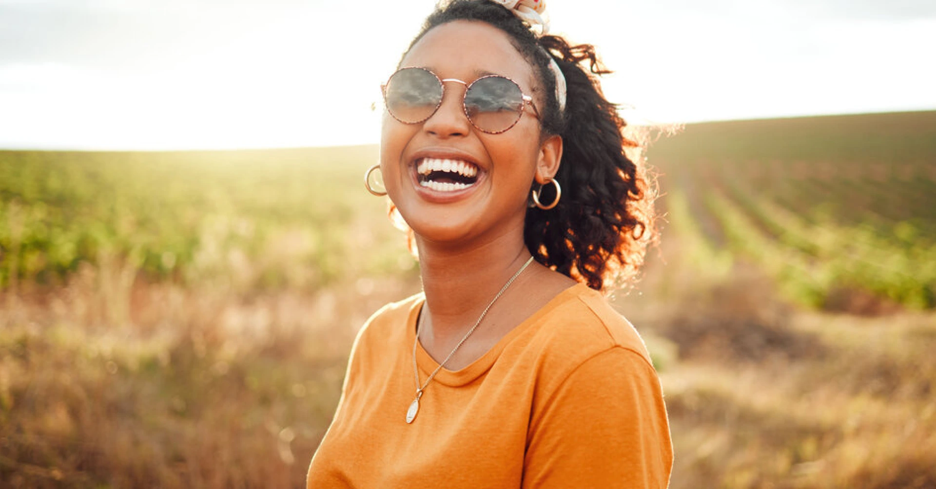 A person with curly hair and wearing round sunglasses and an orange shirt is smiling brightly while standing in front of a sunlit field. The background is slightly blurred, emphasizing the person's joyful expression.